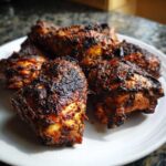 Close-up of several pieces of dark, richly seasoned Jamaican jerk chicken piled on a white plate.