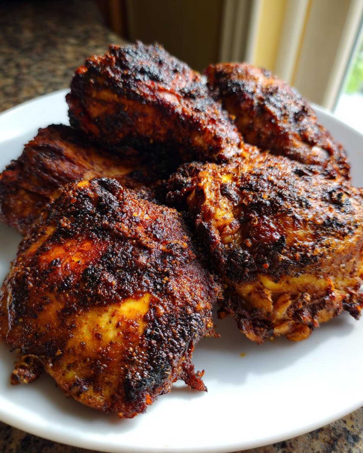 Close-up of several pieces of richly seasoned Jamaican jerk chicken with a dark, charred crust piled on a white plate.