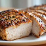 Close-up of juicy baked chicken breast, sliced to show the moist interior and herb-crusted exterior.
