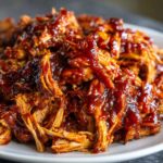 Close-up shot of shredded, tender bbq chicken drenched in a thick, glossy, dark red barbecue sauce on a white plate.