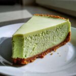 A perfect slice of vibrant green key lime cheesecake with a thick graham cracker crust served on a white plate.