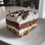A perfect square slice of layered ice cream cake featuring a graham cracker crust, white and chocolate mousse layers, topped with whipped cream and mini chocolate chips.