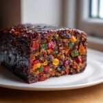 A close-up of a rich, dark, and moist fruit cake recipe packed with colorful candied fruits, sitting on a white plate.