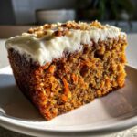 Close-up of a slice of Moist Homemade Carrot Cake topped with thick cream cheese frosting and chopped nuts.