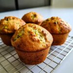 Four golden brown pistachio muffins cooling on a silver wire rack, showcasing visible green pistachio pieces.