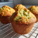 Close-up of four freshly baked, golden-brown pistachio muffins cooling on a wire rack, showing green pistachio pieces.