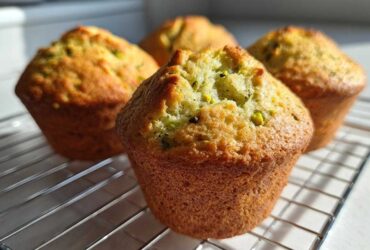 Close-up of four freshly baked, golden-brown pistachio muffins cooling on a wire rack, showing green pistachio pieces.