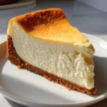 A thick, creamy slice of baked new york cheesecake with a golden-brown top and graham cracker crust, served on a white plate.