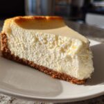 A thick, creamy slice of new york cheesecake resting on a white plate, showing a golden-brown crust.