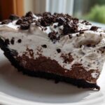 A close-up slice of no bake oreo pie showing a dark crust, chocolate filling, and thick white whipped topping sprinkled with cookie crumbs.
