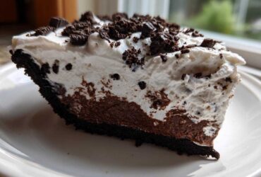 A close-up slice of no bake oreo pie showing a dark crust, chocolate filling, and thick white whipped topping sprinkled with cookie crumbs.
