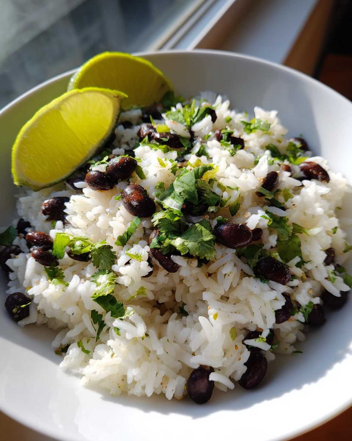 A white bowl filled with fluffy white rice mixed with black beans and fresh cilantro, garnished with two lime wedges, showcasing a simple beans and rice dish.