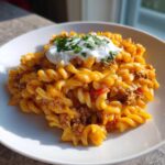 A close-up serving of cheesy taco pasta with ground beef, topped with sour cream and fresh cilantro.