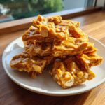 A stack of golden, glossy pieces of homemade peanut brittle recipe resting on a small white plate.