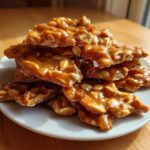 A stack of golden brown, glossy pieces of homemade peanut brittle recipe resting on a white plate.