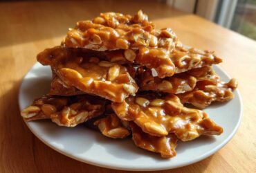A stack of golden brown, glossy pieces of homemade peanut brittle recipe resting on a white plate.