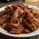 A close-up of shredded, tender pioneer woman slow cooker pulled pork coated in rich barbecue sauce on a white plate.