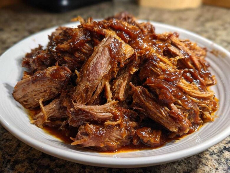 A close-up of shredded, tender pioneer woman slow cooker pulled pork coated in rich barbecue sauce on a white plate.