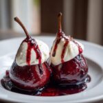 Close-up of two decadent poached pears, deep red from poaching liquid, topped with whipped cream and berry syrup.