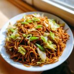 A close-up of a white plate piled high with savory brown fried noodles mixed with sautéed cabbage and topped with fresh green onions.