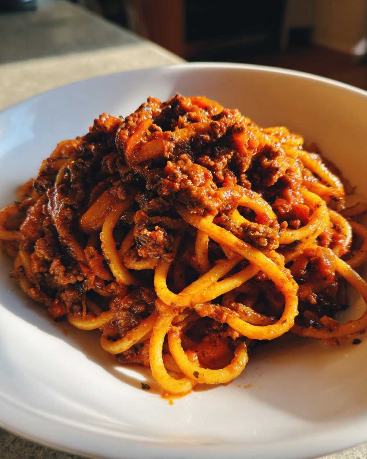 Close-up of thick spaghetti noodles coated generously with rich, meaty bolognese sauce in a white bowl.