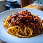A close-up of spaghetti topped generously with rich, dark Bolognese sauce on a white plate.