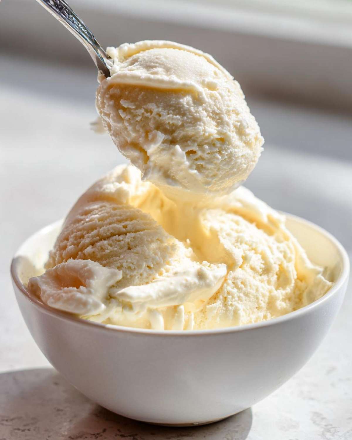 A spoonful of creamy, pale yellow banana ice cream being lifted from a white bowl filled with more banana ice cream.