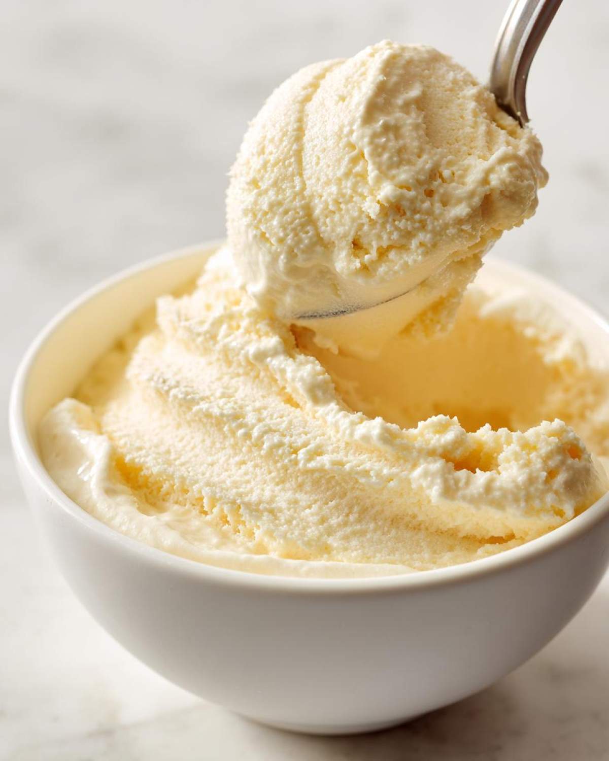 A metal spoon scoops a perfect ball of creamy, pale yellow banana ice cream from a white bowl.