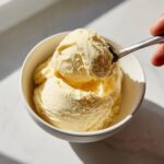 A scoop of creamy, pale yellow banana ice cream being lifted from a white bowl with a metal spoon.