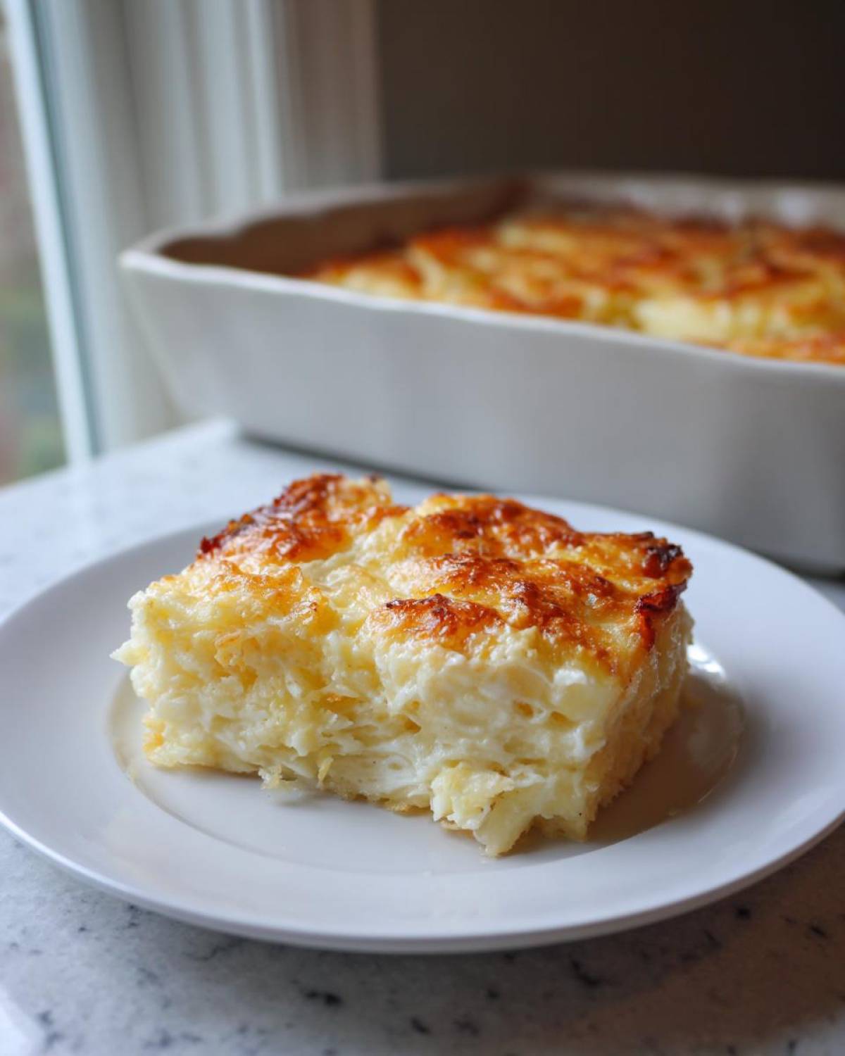 A square serving of creamy funeral potatoes recipe with a golden, bubbly cheese topping on a white plate.