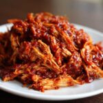Close-up of tender, shredded bbq chicken coated generously in rich, dark red barbecue sauce, served on a white plate.