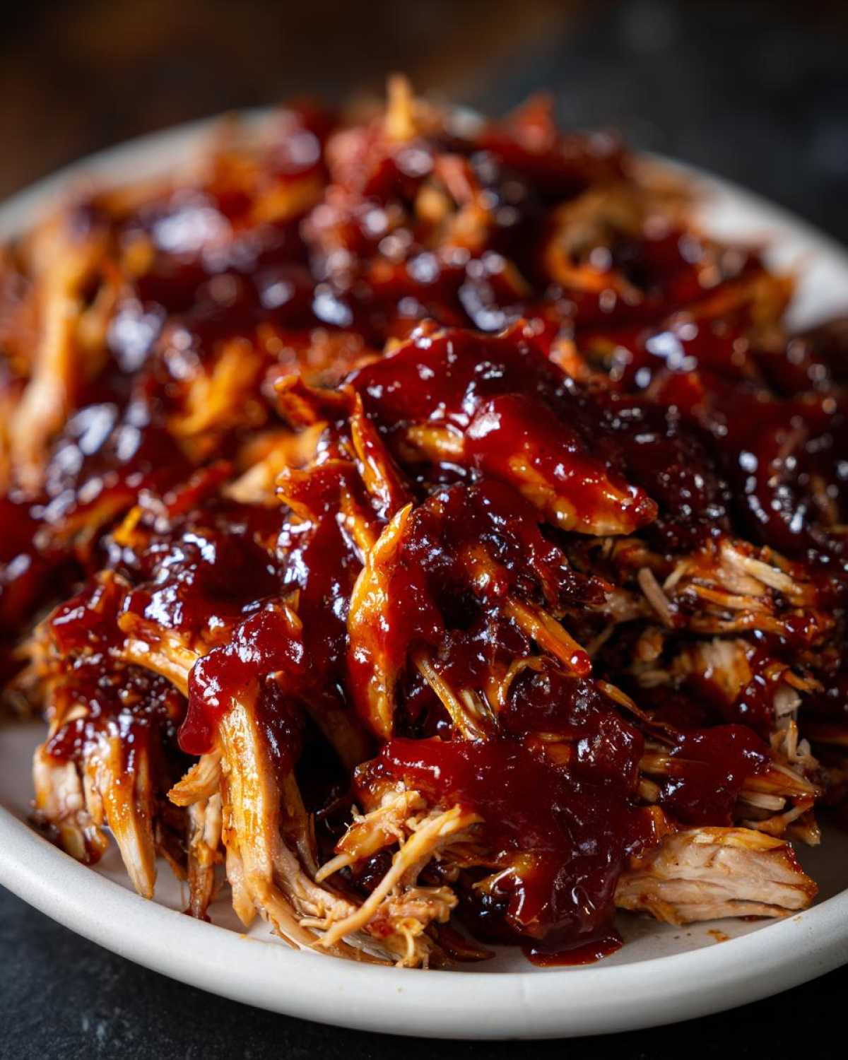 Close-up of juicy shredded bbq chicken smothered in thick, glossy barbecue sauce on a white plate.
