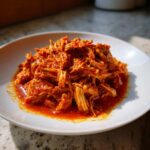 A close-up of shredded chicken tinga coated in a vibrant, rich red sauce, served on a white plate.