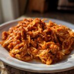 A close-up of shredded chicken tinga, coated in a vibrant orange-red sauce, served on a white plate.