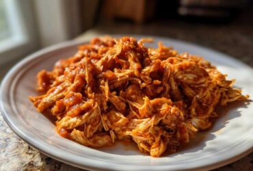 A close-up of shredded chicken tinga, coated in a vibrant orange-red sauce, served on a white plate.