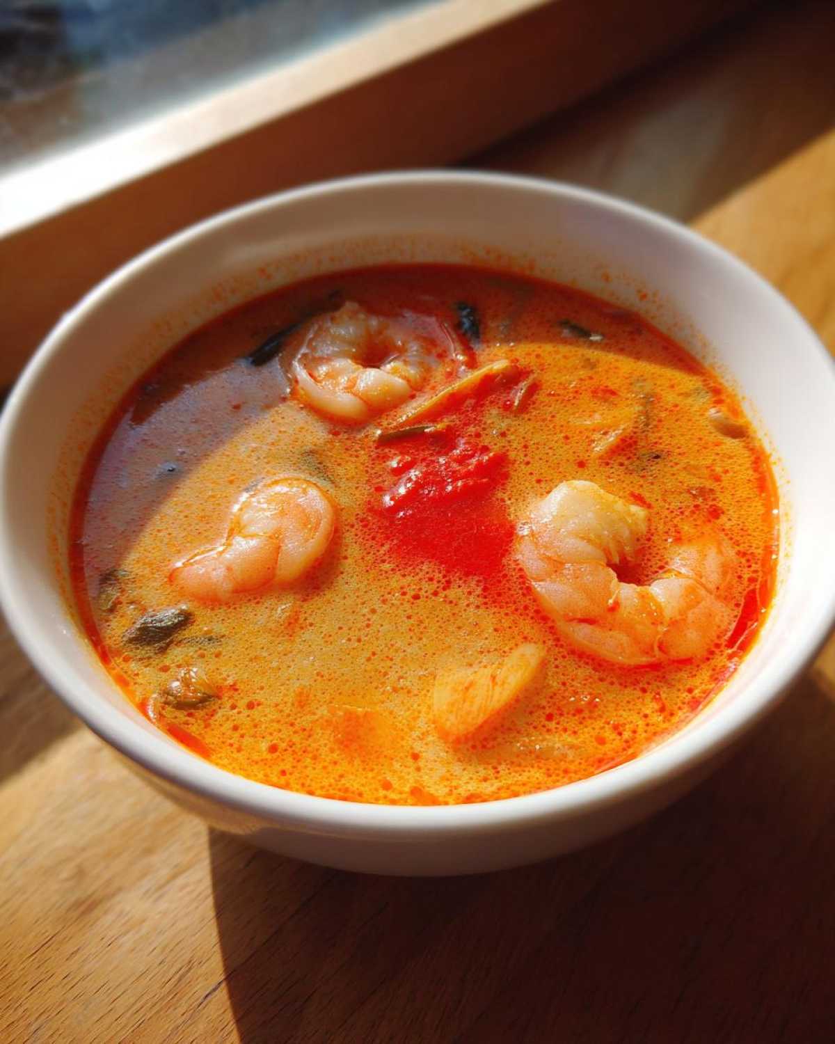 Close-up of a white bowl filled with bright orange and red tom yum soup featuring plump shrimp.