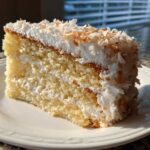 Close-up of a moist slice of 3 layer coconut cake recipe, topped with white frosting and toasted coconut flakes.
