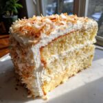 A moist slice of layered coconut cake covered in white frosting and topped with toasted coconut flakes.