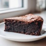 A close-up of a dense, dark slice of flourless chocolate cake dusted with powdered sugar on a white plate.
