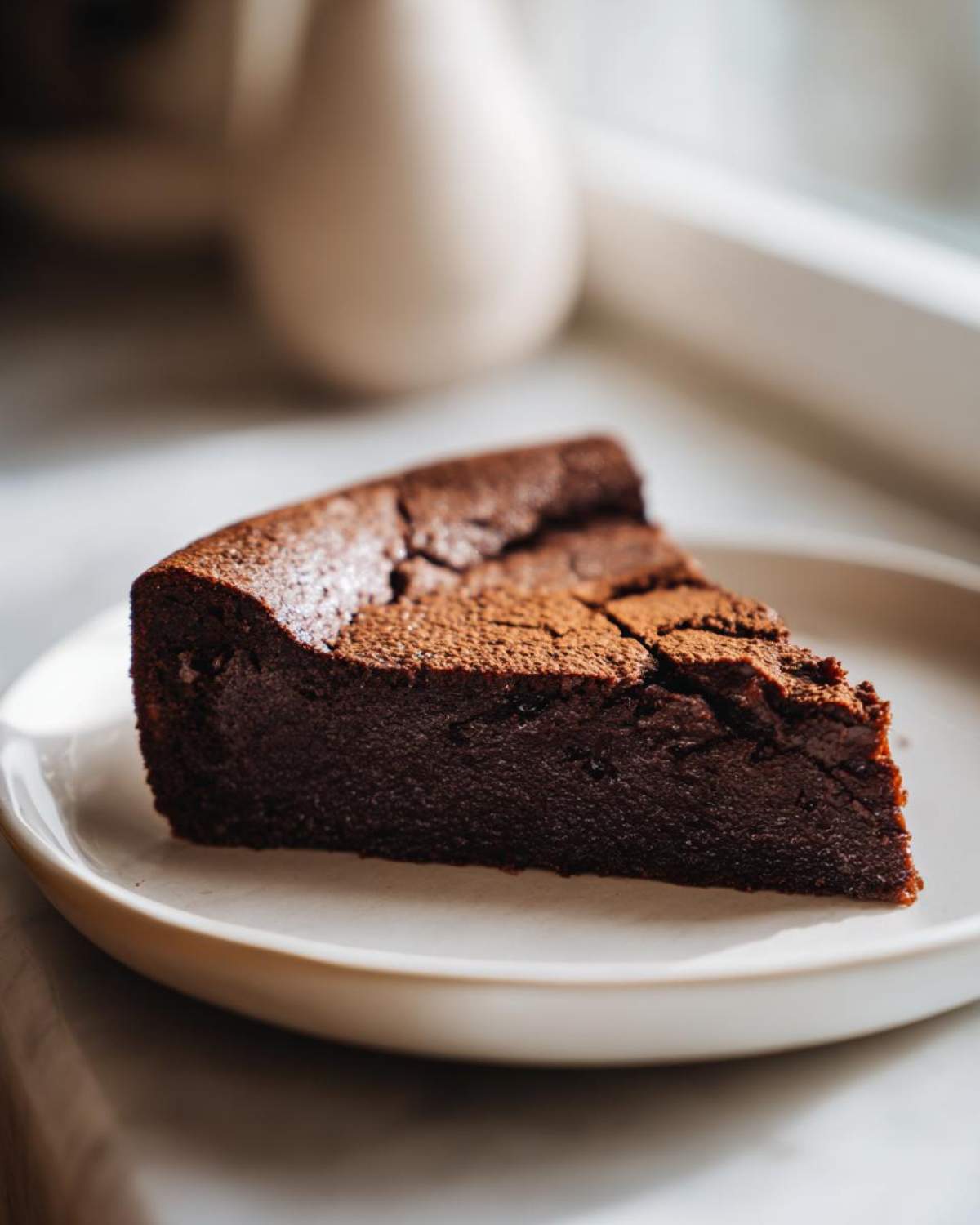A decadent slice of flourless chocolate cake dusted with cocoa powder, served on a light ceramic plate.