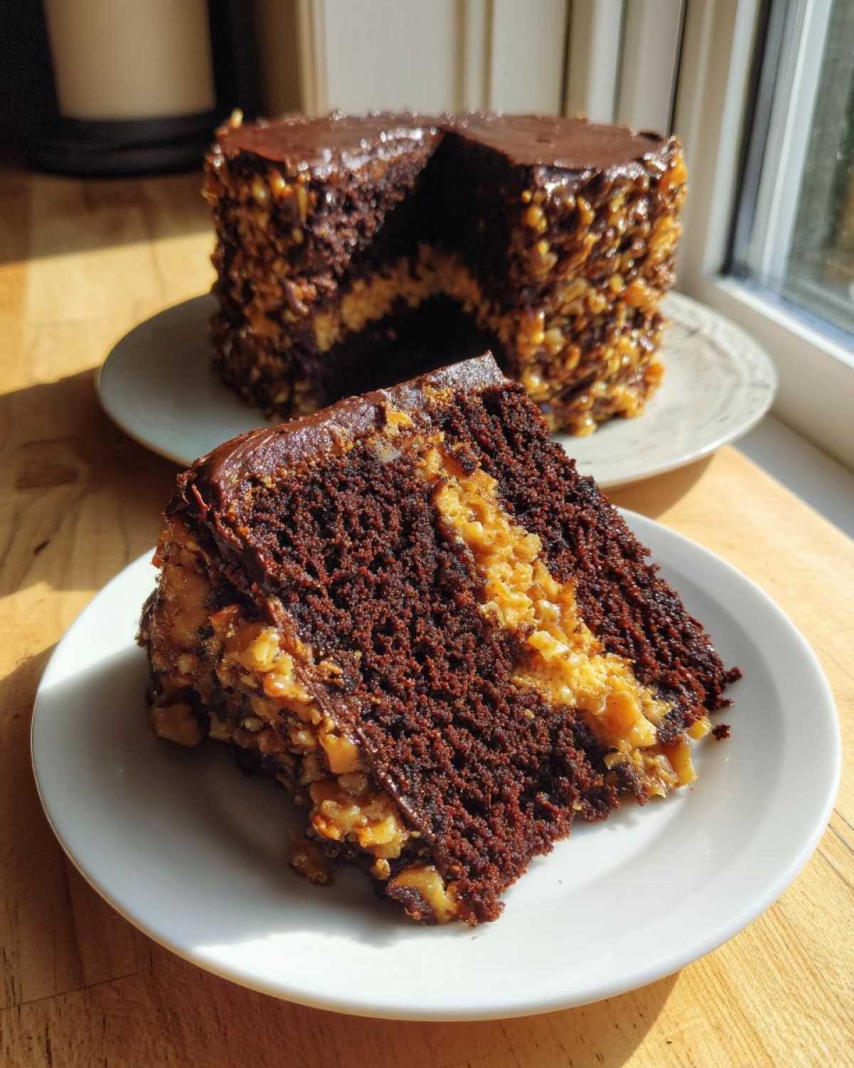 A slice of moist german chocolate cake showing dark layers and coconut-pecan filling, with the rest of the cake in the background.