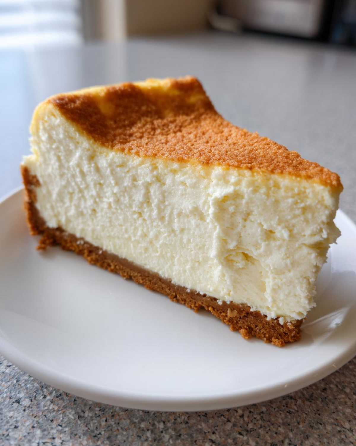 A thick slice of creamy new york cheesecake with a graham cracker crust and a lightly browned top, served on a white plate.