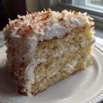 A moist slice of layered coconut cake with white frosting and toasted coconut flakes on top.