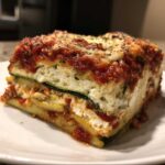A close-up of a thick, layered slice of zucchini lasagna showing zucchini noodles, ricotta cheese, and rich marinara sauce.