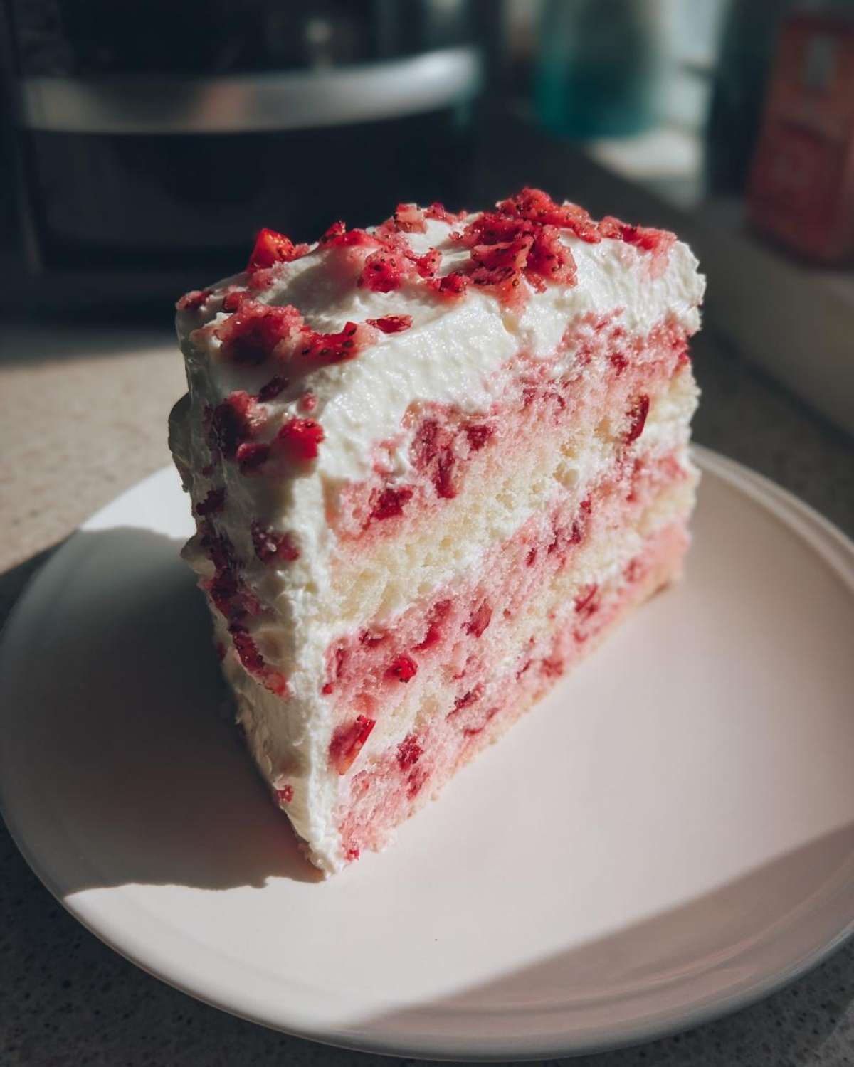 A tall slice of layered strawberry cake with white frosting and crushed strawberry topping, sitting on a white plate.