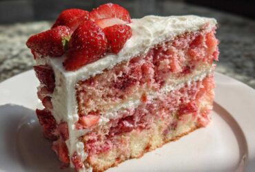 A moist, layered slice of strawberry cake with white frosting and topped with fresh sliced strawberries.