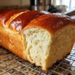 A freshly baked, golden-brown Japanese milk bread loaf with a slice cut off, showing its fluffy interior, cooling on a wire rack.