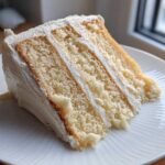 Close-up of a moist, three-layer slice of vanilla cake recipe with thick white frosting.