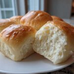 Close-up of soft, golden-topped sourdough discard rolls on a white plate, one roll broken open showing the fluffy interior.