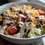 A close-up of a white bowl filled with a vibrant southwest salad featuring shredded chicken, black beans, corn, tomatoes, and creamy dressing.